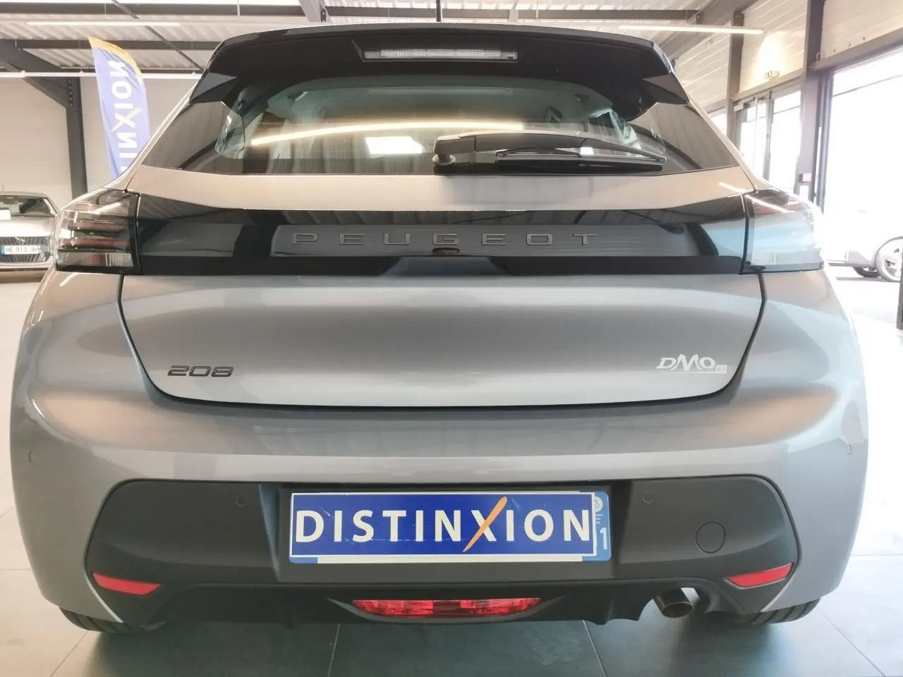 Vue arrière d'une Peugeot 208 2024 gris Artense avec logo distinctif et plaque d'immatriculation Distinxion en intérieur showroom.