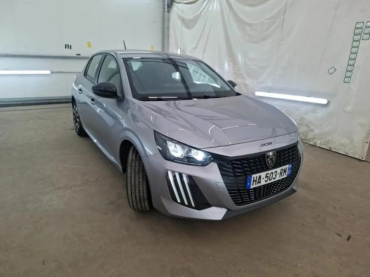 Peugeot 208 gris clair vue 3/4 avant droit, phares allumés et calandre noire distinctive, dans un garage.