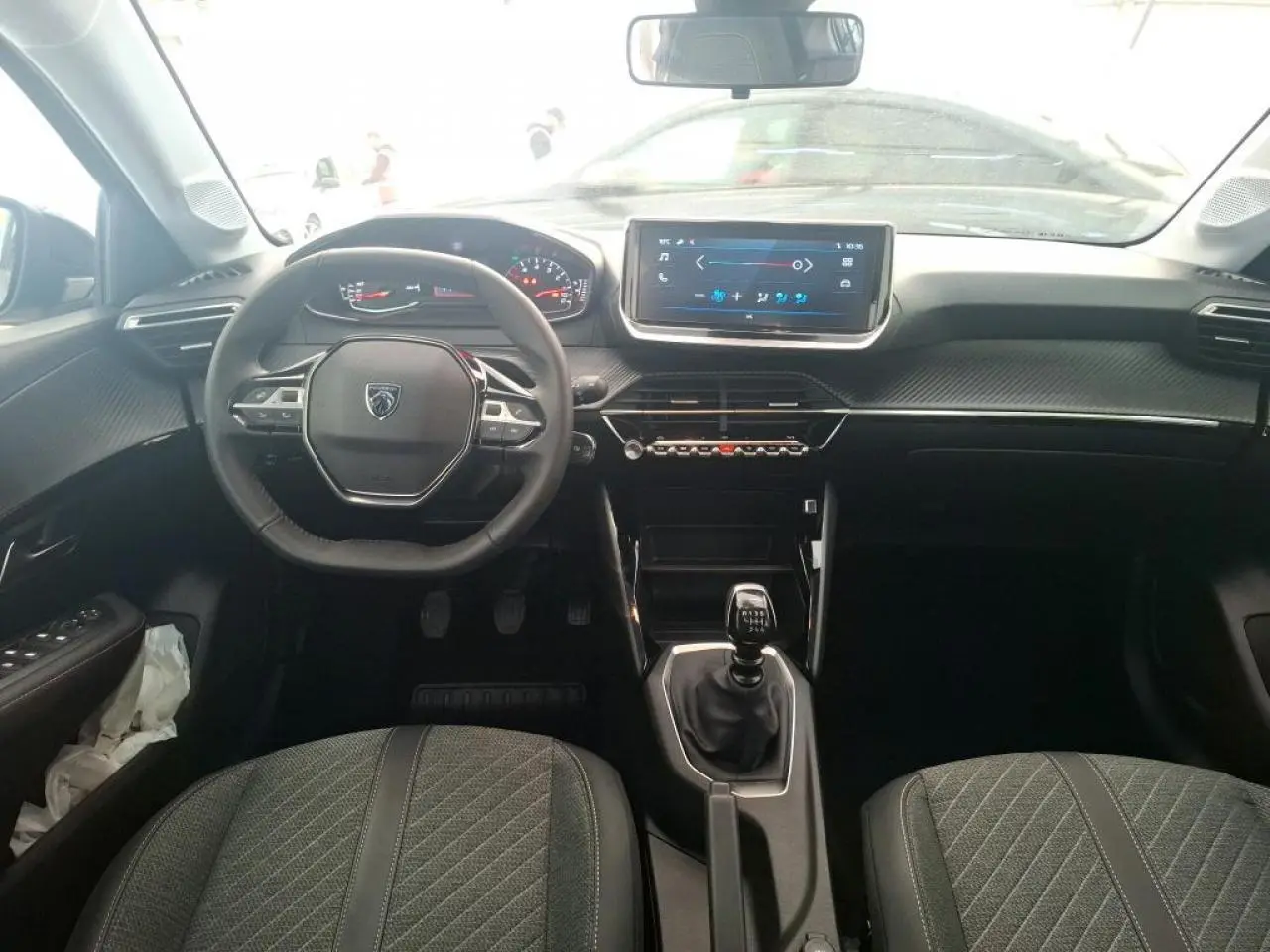 Vue intérieure frontale du tableau de bord et volant de la Peugeot 208 2024, avec boîte manuelle et sièges gris.