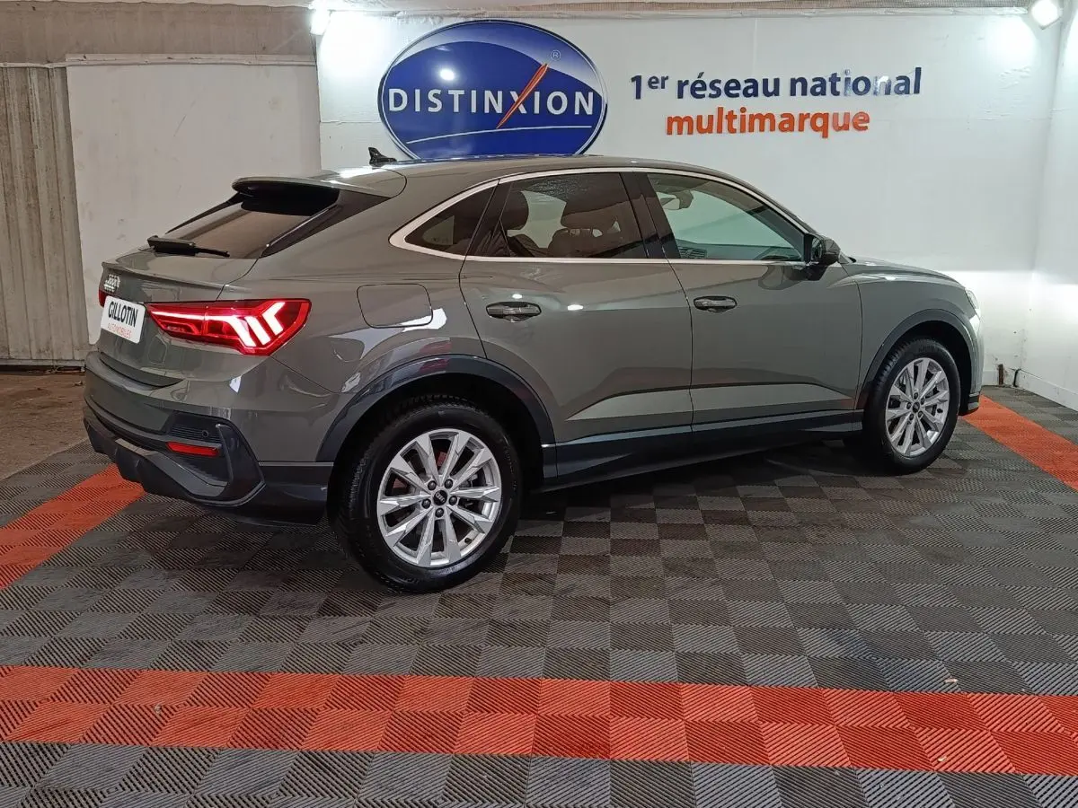 Audi Q3 Sportback gris foncé vue 3/4 arrière droit, avec feux arrière allumés dans un showroom.
