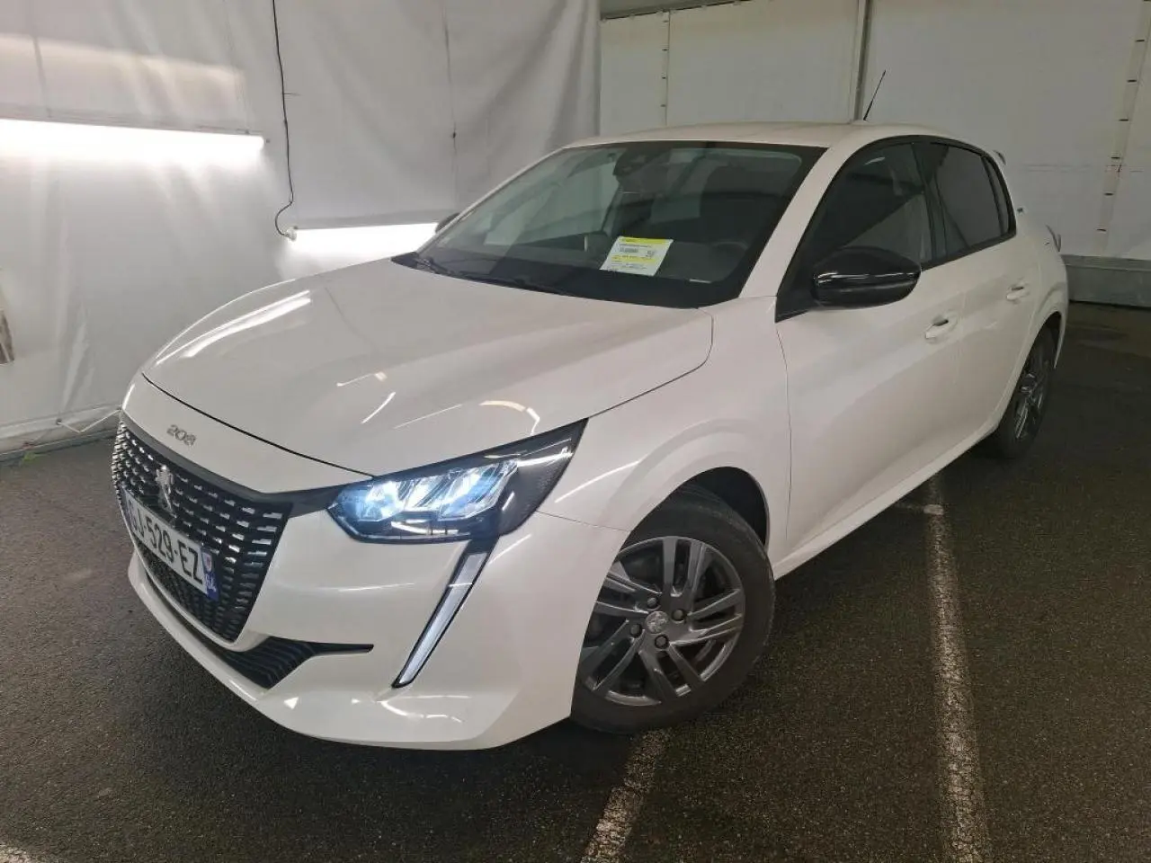 Peugeot 208 blanche vue en 3/4 avant droit, phares LED allumés et jantes alliage 16 pouces visibles.