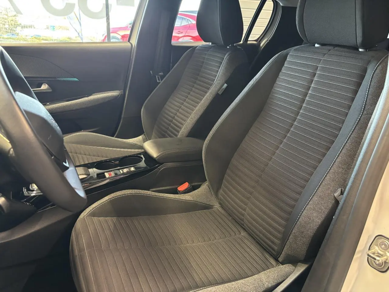 Vue intérieure côté avant droit montrant les sièges tissu gris et accoudoir central de la Peugeot 208 blanche.