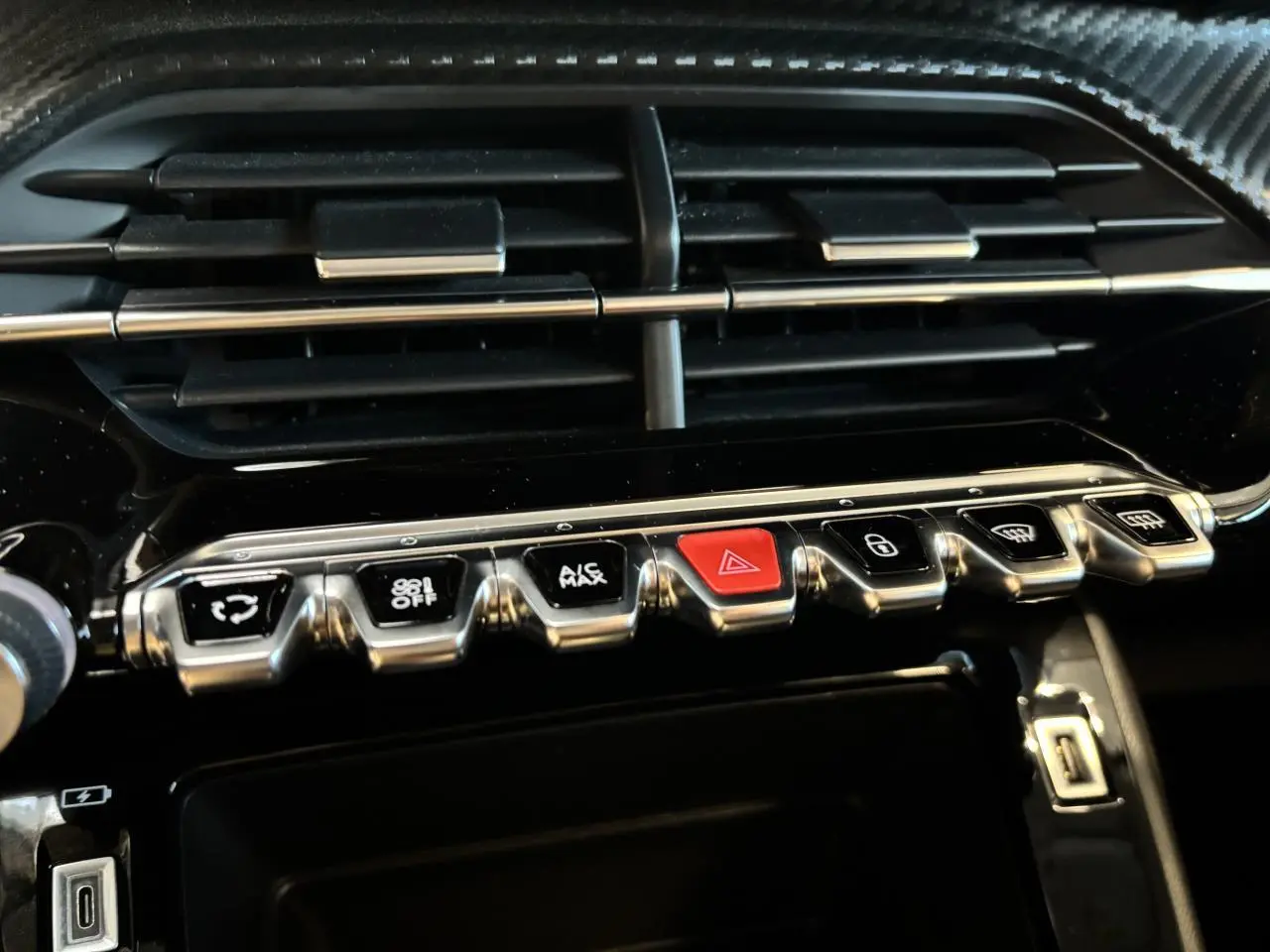 Gros plan sur la rangée de boutons chromés et noirs avec bouton rouge d’alerte, tableau de bord noir brillant Peugeot 208 blanc.