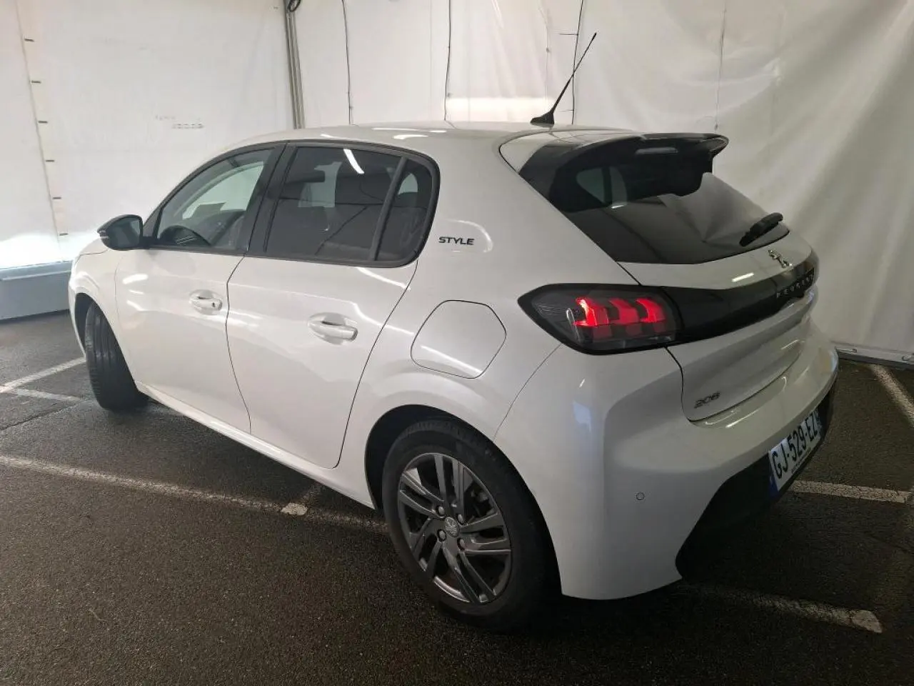 Vue 3/4 arrière droite d'une Peugeot 208 blanche avec jantes alliage et bandeau noir brillant entre feux arrière.