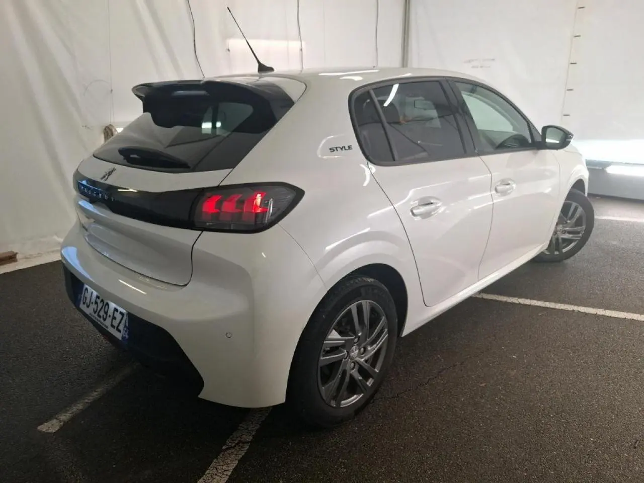 Peugeot 208 blanche vue 3/4 arrière droit, avec bandeau noir brillant entre feux et jantes alliage 16 pouces.