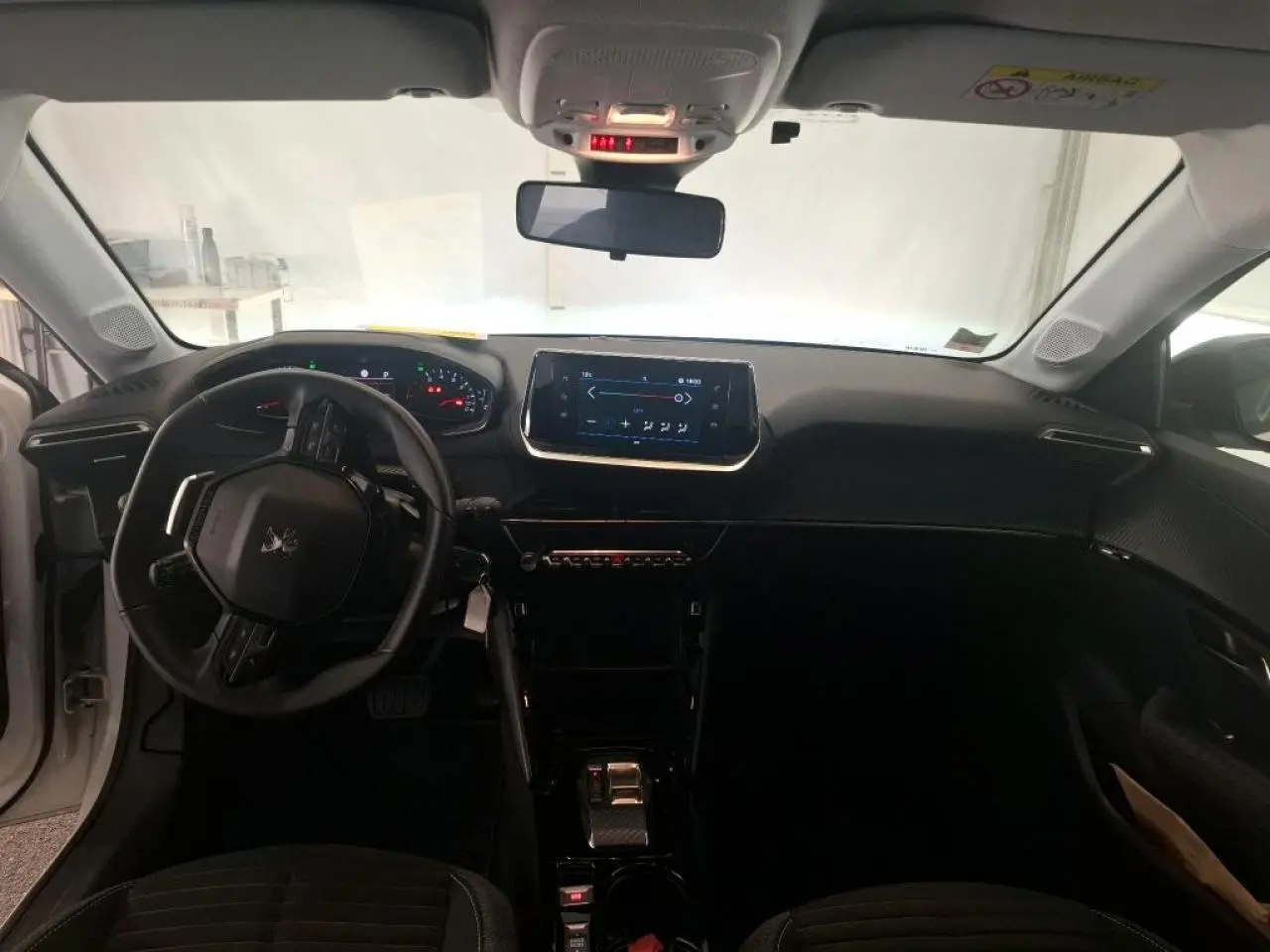 Vue intérieure avant de la Peugeot 208 blanche, montrant le tableau de bord noir avec écran tactile et volant compact cuir.