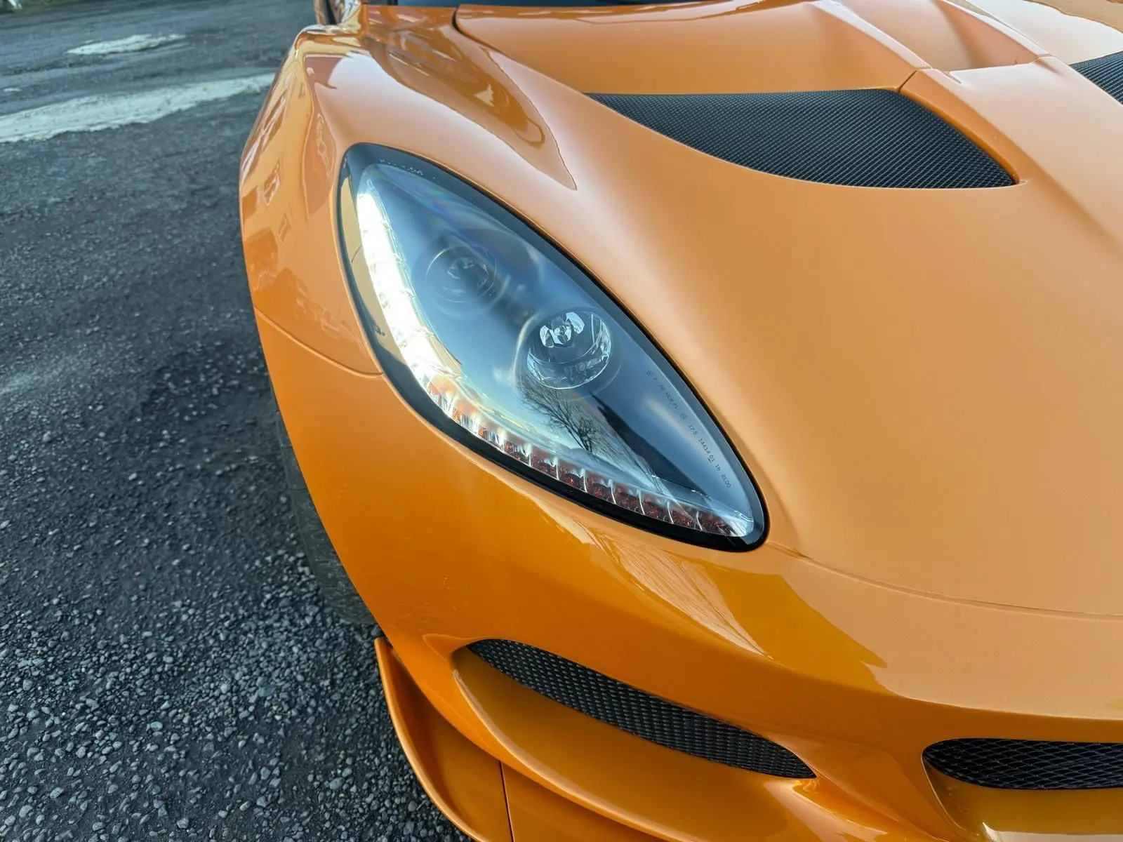 Gros plan sur l'avant gauche orange vif de la Lotus Elise 3 Cup 250 2021 avec phare halogène et prise d'air en carbone.
