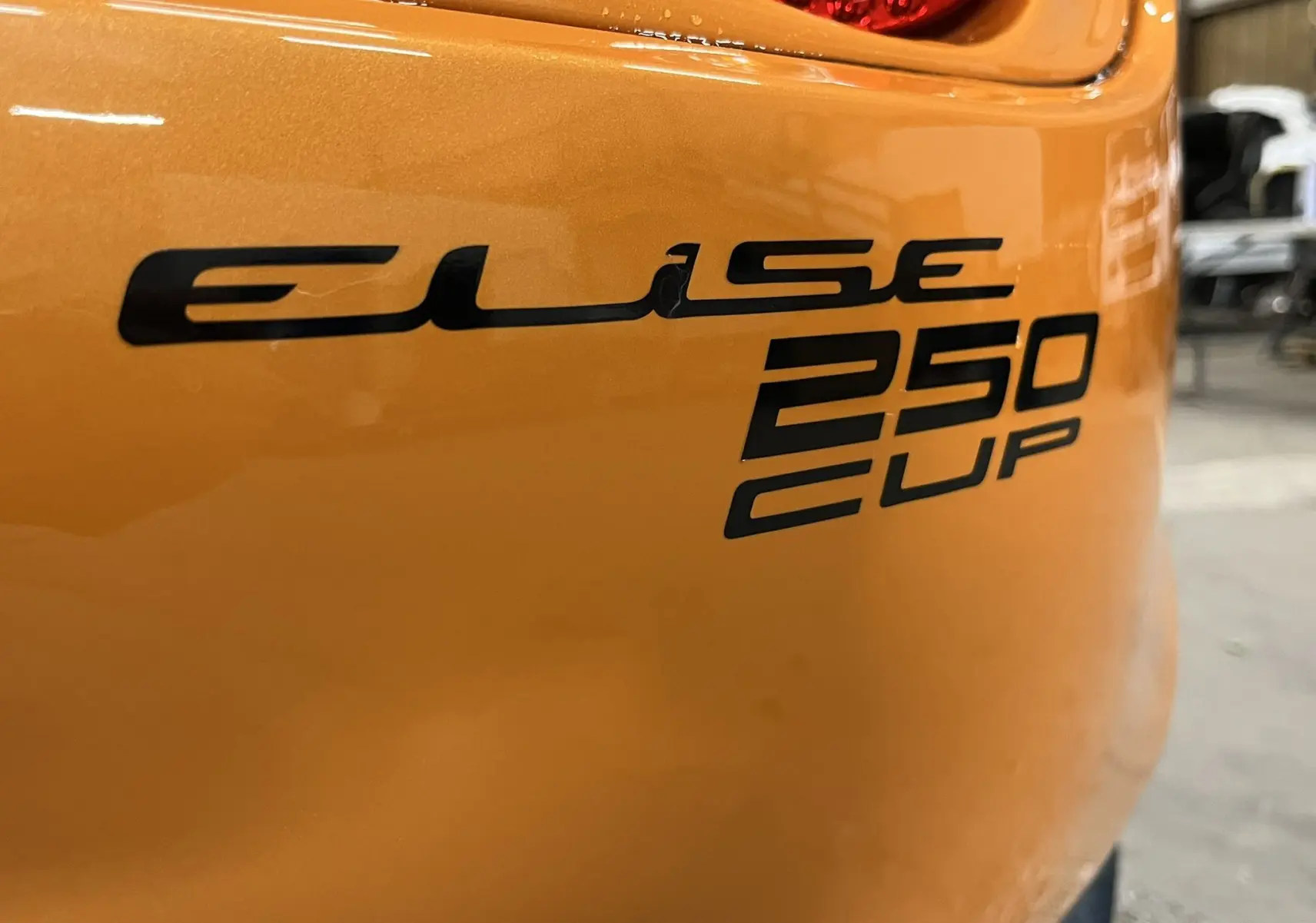 Gros plan sur l'arrière orange de la Lotus Elise 3 Cup 250 2021 avec logo noir et feu arrière partiel visible.