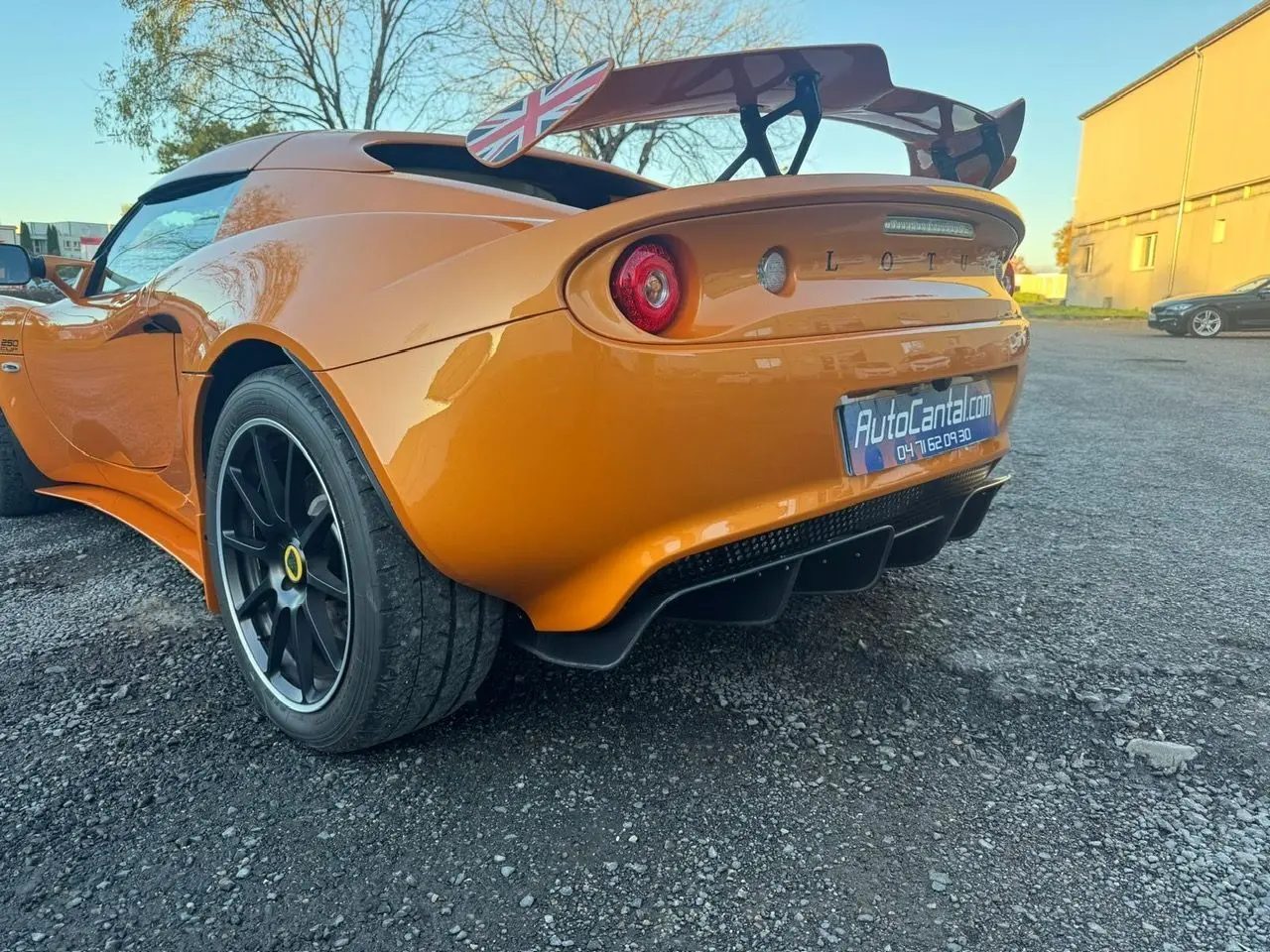 Vue 3/4 arrière droite de la Lotus Elise 3 Cup 250 orange avec jantes noires satinées et aileron arrière marqué du drapeau britannique.