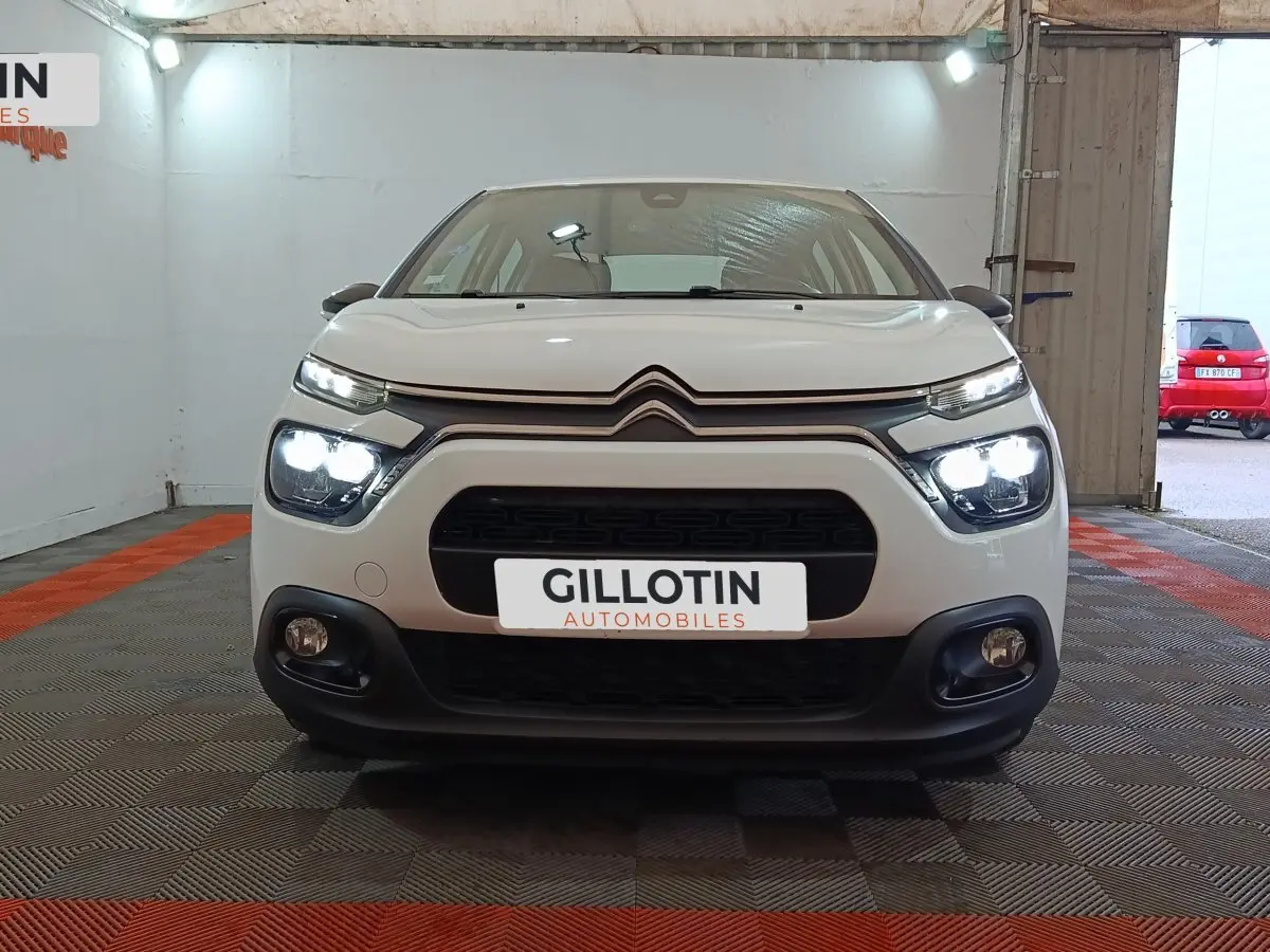 Vue de face d'une Citroën C3 blanche avec phares allumés dans un garage au sol carrelé rouge et gris.