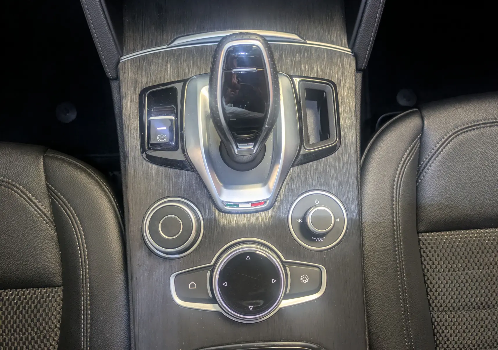 Vue plongeante sur la console centrale noire en bois de l'Alfa Romeo Stelvio 2023 avec levier de vitesse et commandes circulaires.