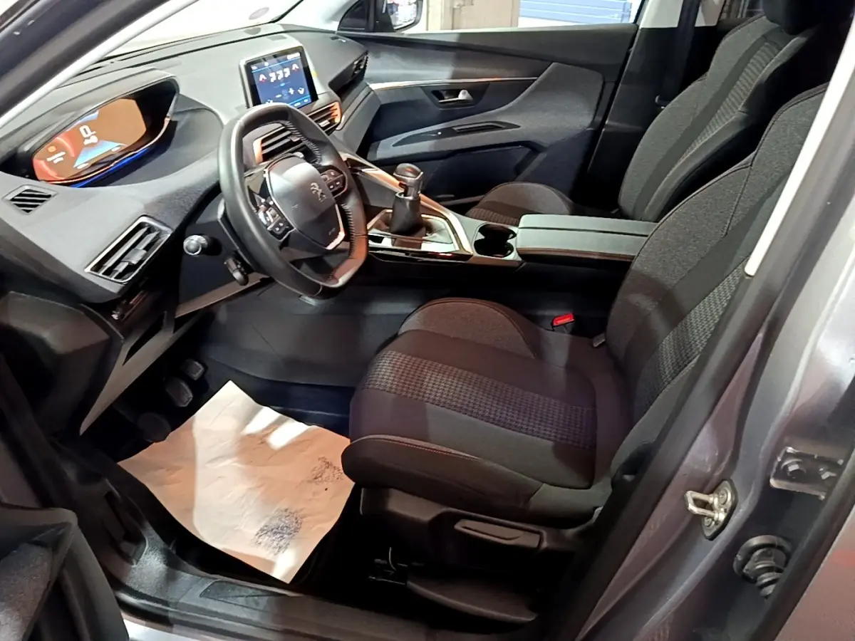 Vue intérieure côté conducteur du Peugeot 3008 gris clair, montrant le volant cuir, tableau de bord digital et levier de vitesse manuel.