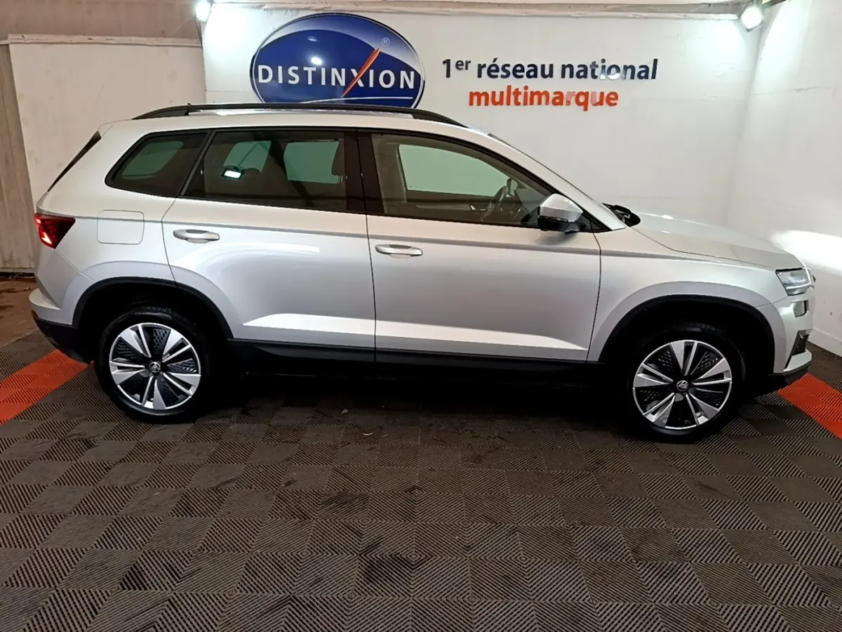 Vue latérale droite d’un Skoda Karoq gris clair 2022 avec jantes alliage et barres de toit noires en showroom.