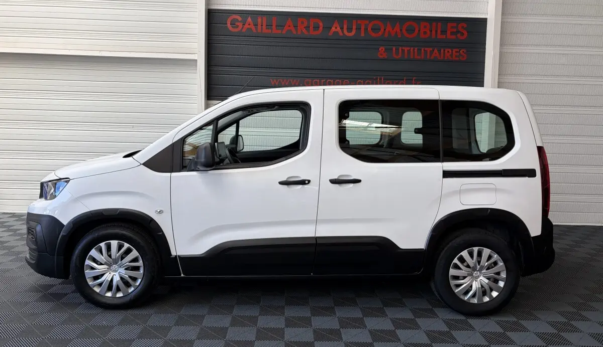 Profil droit d'un Peugeot Rifter blanc 2021 avec protections noires sur bas de caisse et jantes argentées, stationné en intérieur.