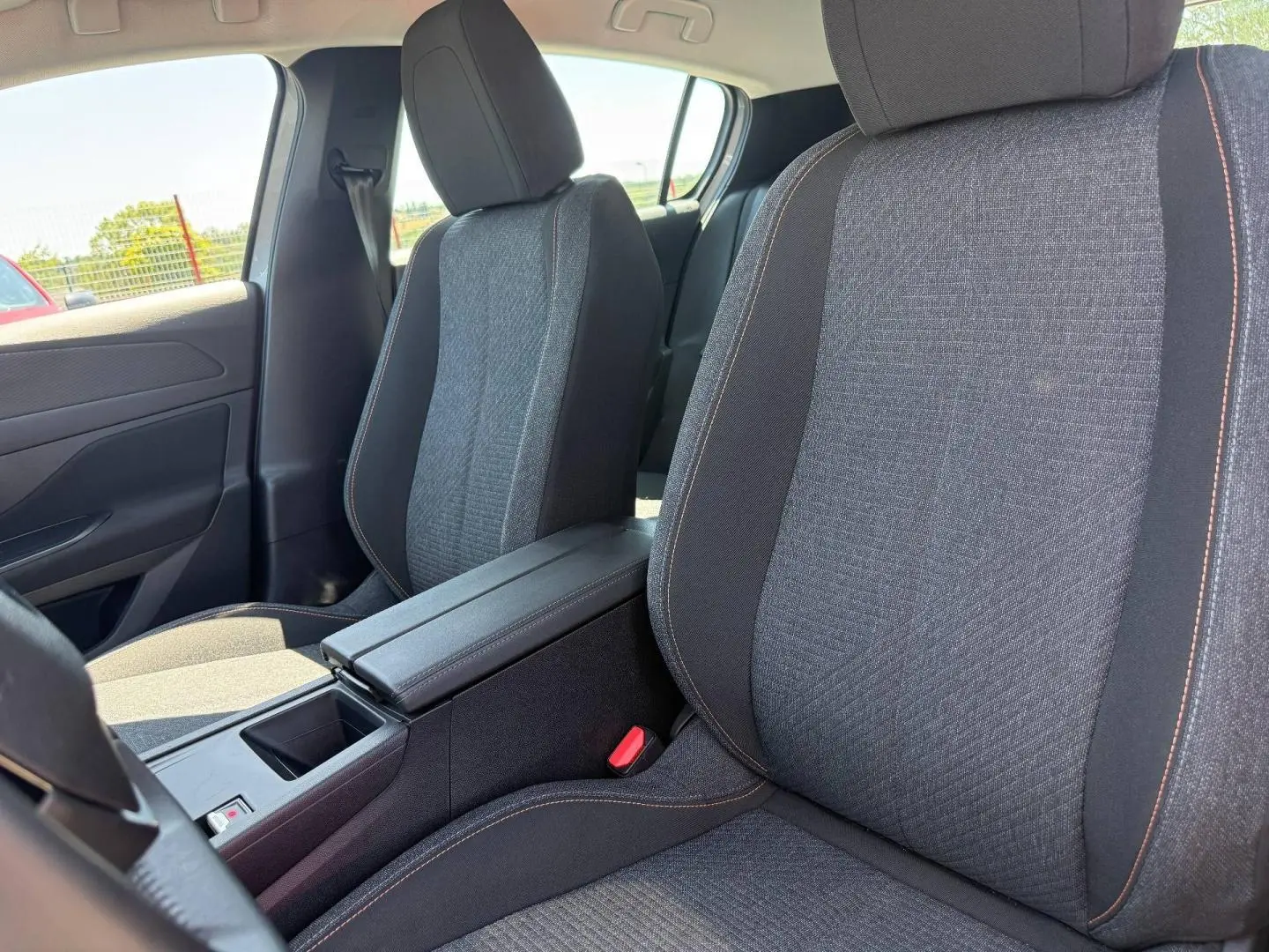 Vue intérieure rapprochée des sièges avant gris foncé avec surpiqûres orange et accoudoir central ouvert d'une Peugeot e-308 noire.