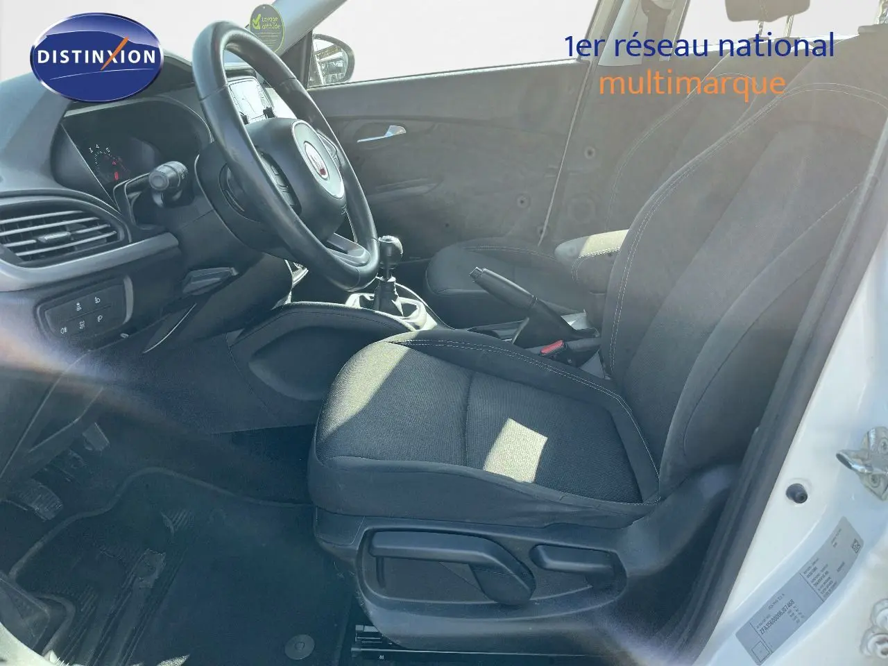 Vue intérieure côté conducteur d'une Fiat Tipo blanche 2018, montrant les sièges noirs et la boîte manuelle.