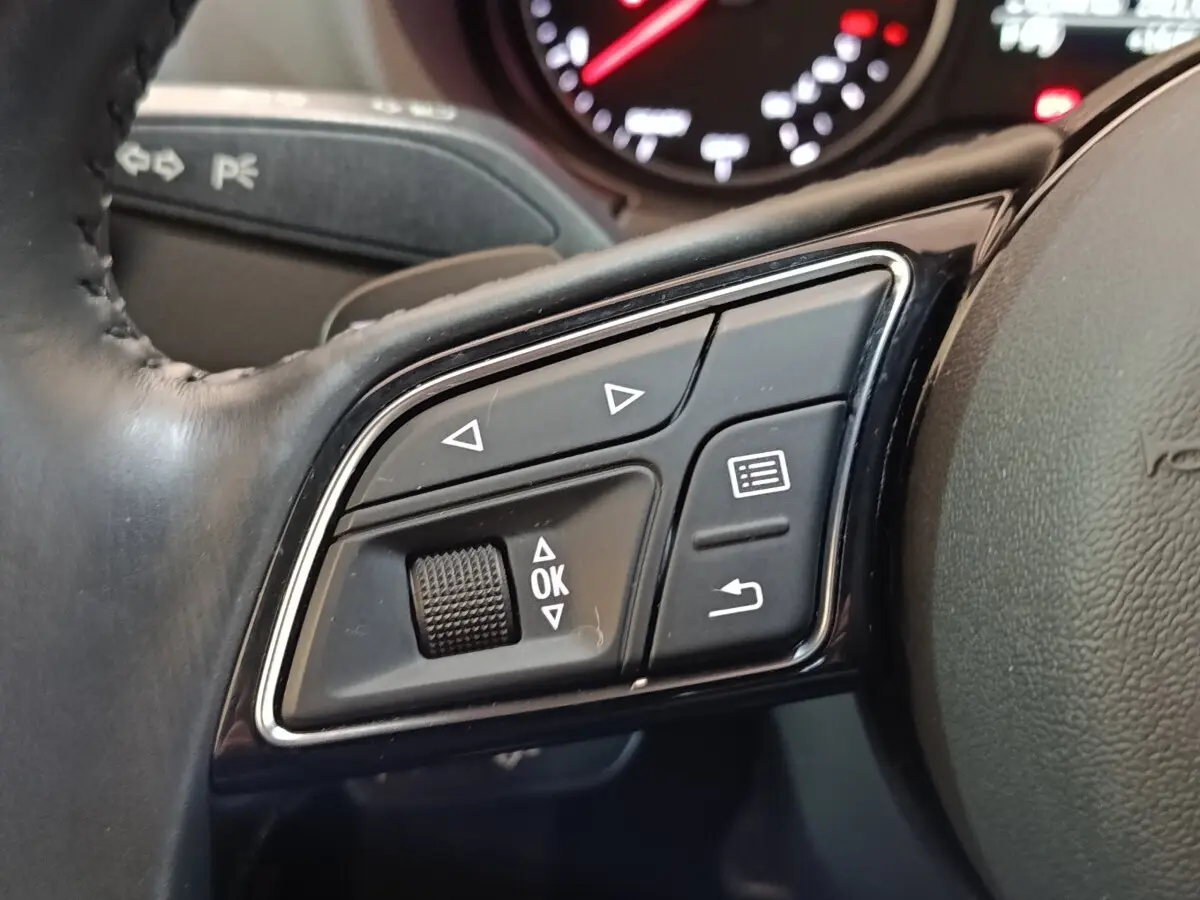 Gros plan sur les commandes noires multifonctions du volant cuir d'une Audi A3 blanche, avec tableau de bord en arrière-plan flou.