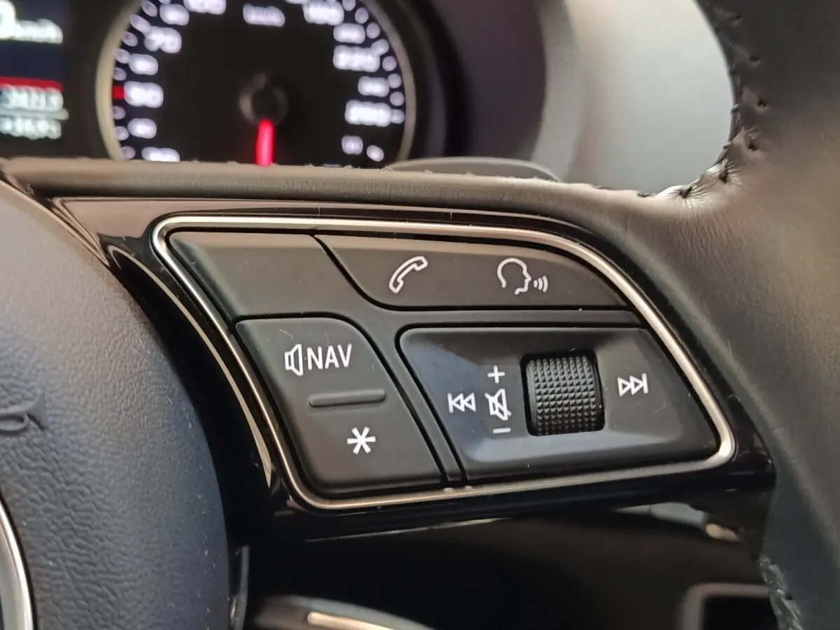 Gros plan sur les commandes multifonctions du volant cuir de l'Audi A3 blanc, avec tableau de bord flou en arrière-plan.