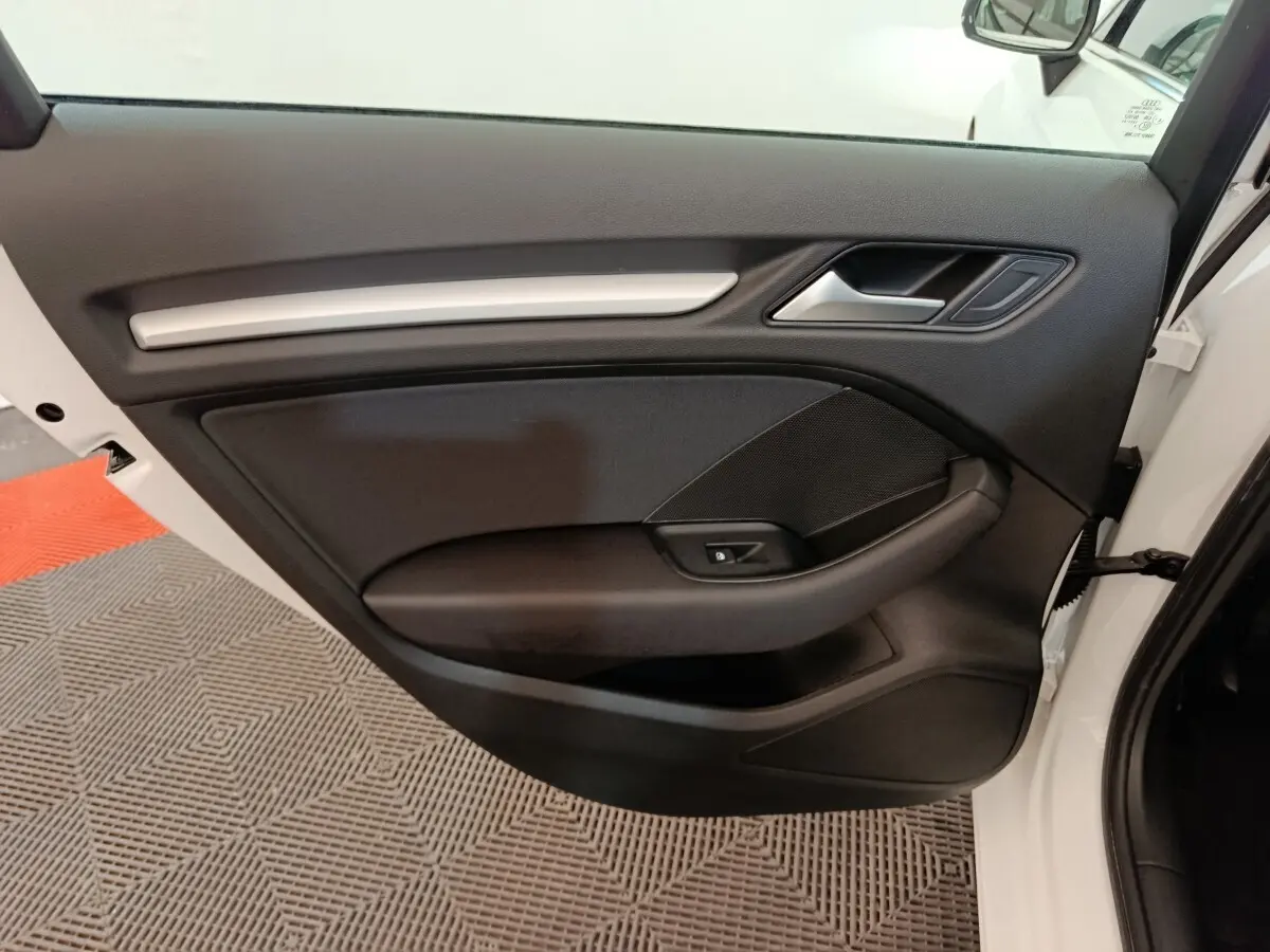 Vue rapprochée de la porte avant droite d’une Audi A3 blanche, montrant la garniture intérieure noire avec poignée et commande de vitre.