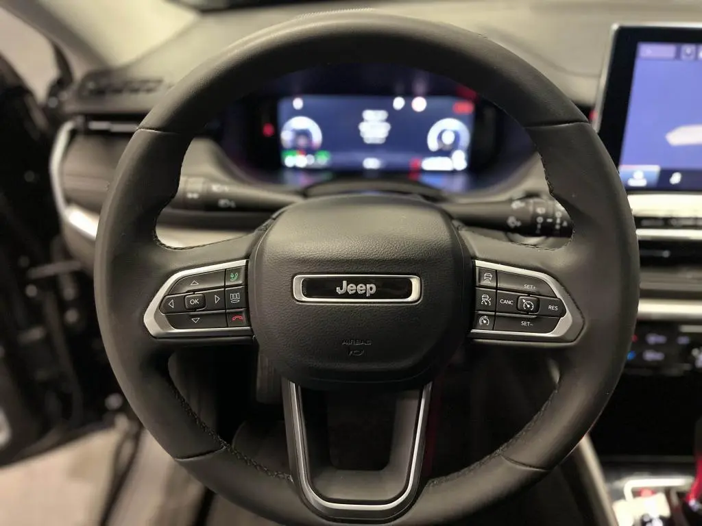 Vue rapprochée du volant noir du Jeep Compass 2024 avec commandes intégrées et tableau de bord numérique flou en arrière-plan.