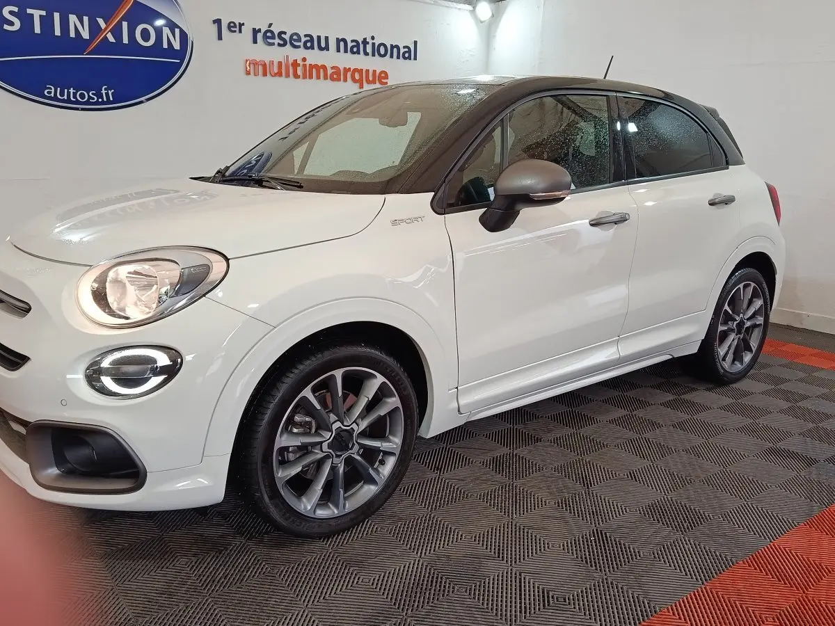 Vue 3/4 avant droit d'une FIAT 500X blanche avec toit noir et jantes alliage, en intérieur sur sol damier.