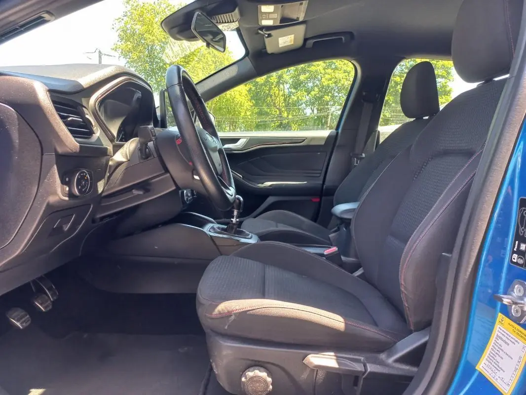 Vue intérieure côté conducteur d'une Ford Focus bleu 2019, montrant les sièges noirs avec surpiqûres rouges et la boîte manuelle.