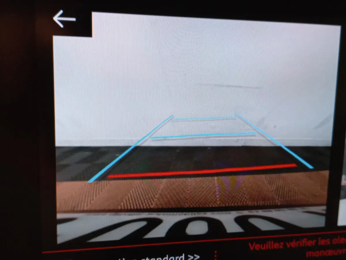 Vue arrière caméra de recul affichant les lignes de guidage bleues et rouges sur un sol sombre, intérieur de Peugeot 208 blanc visible