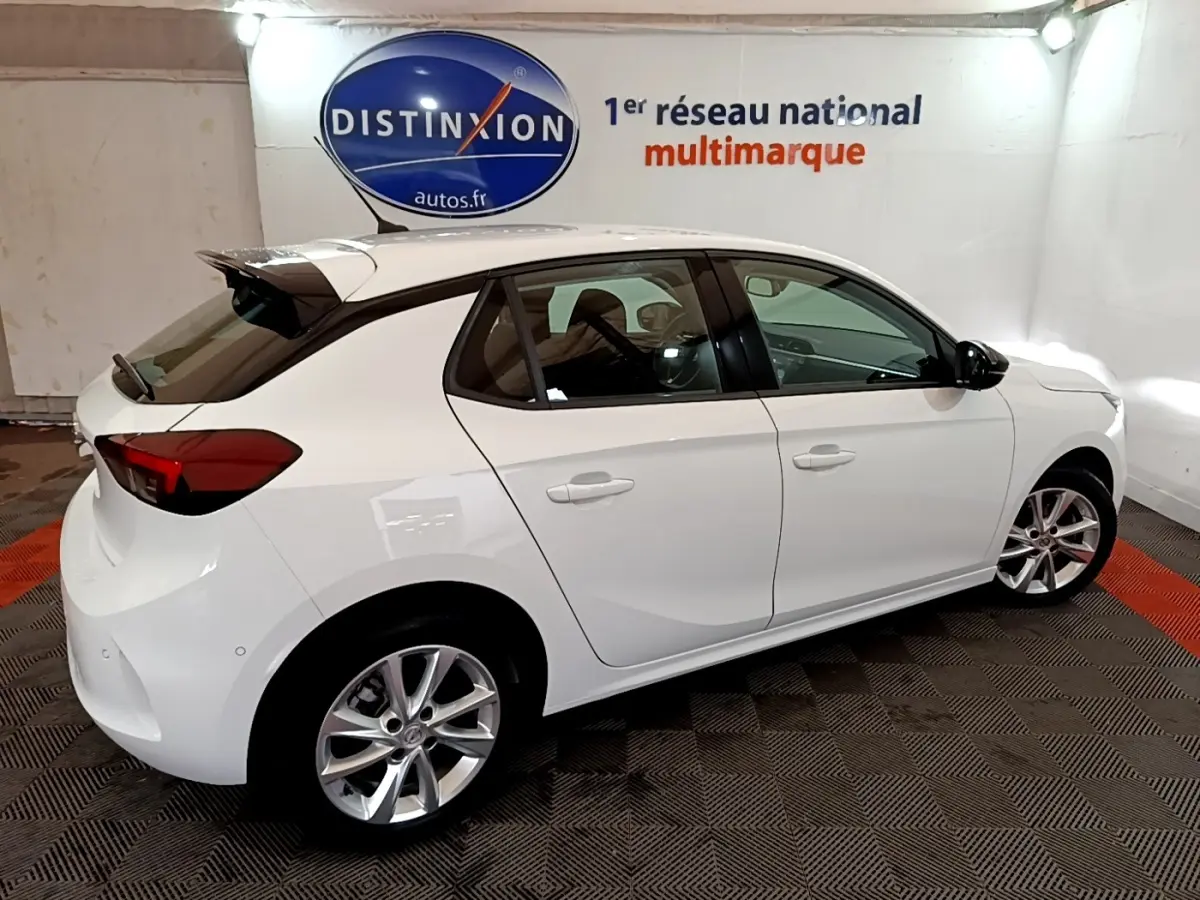 Vue 3/4 arrière droite d'une Opel Corsa blanche 2023 avec jantes alliage et spoiler de toit dans un showroom.