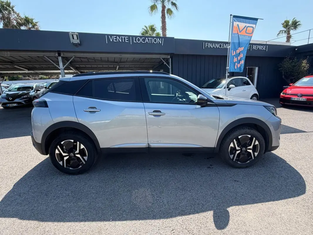 Profil côté gauche d’un Peugeot 2008 Hybrid 145 gris 2025 avec jantes noires et toit noir, stationné en extérieur.