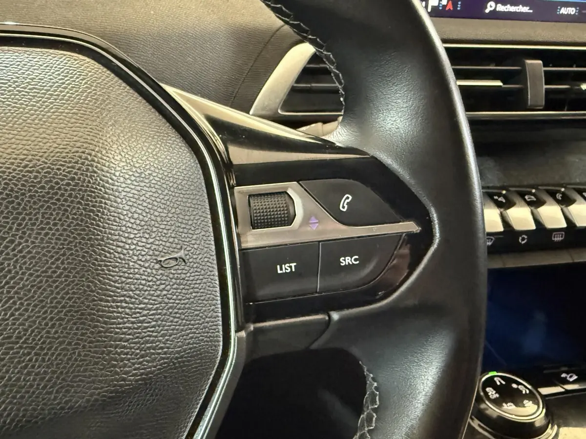 Gros plan sur le volant cuir noir du Peugeot 3008 Puretech 130, avec commandes audio et tableau de bord en arrière-plan.