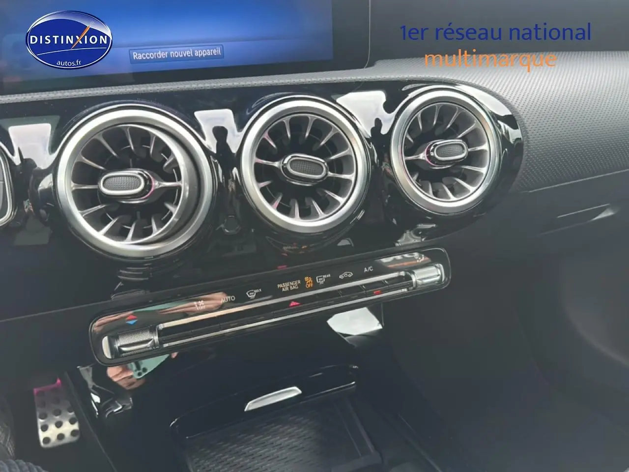 Gros plan sur la console centrale noire brillante avec trois aérateurs ronds et commandes climatisation d'une Mercedes Classe A bleue.