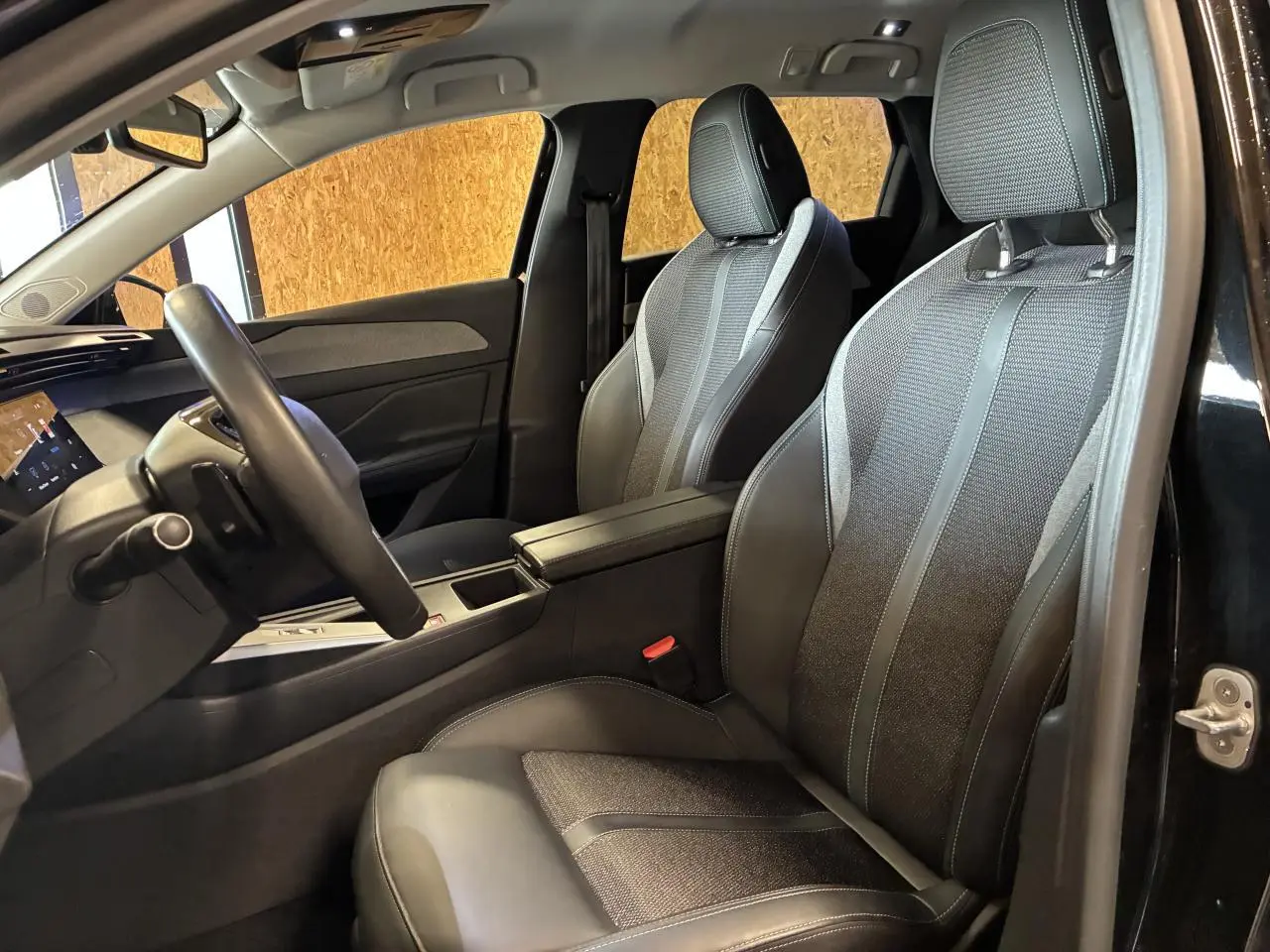 Vue intérieure côté conducteur de la Peugeot 308 SW noire, montrant les sièges avant en tissu et cuir avec accoudoir central.