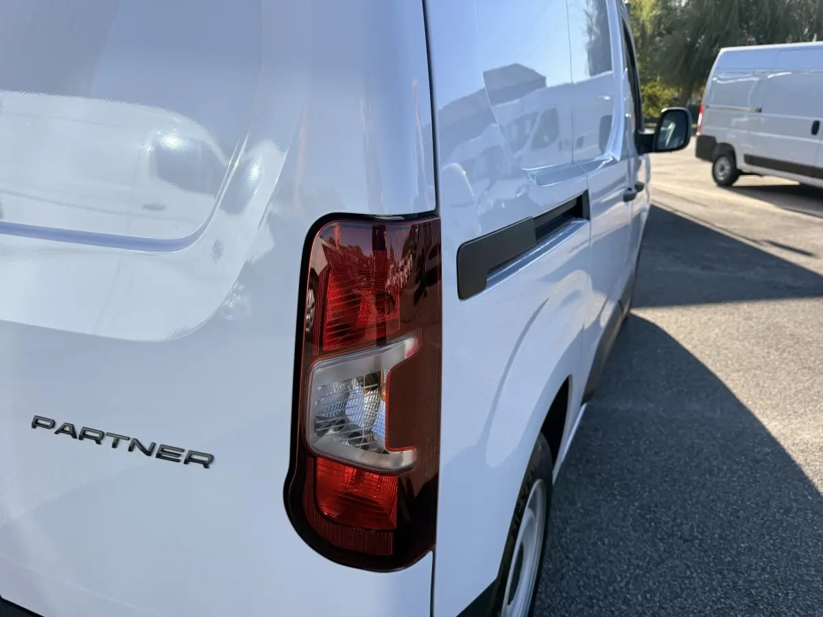 Vue arrière côté droit d’un Peugeot Partner blanc avec feu arrière et logo visible sur une route ensoleillée.