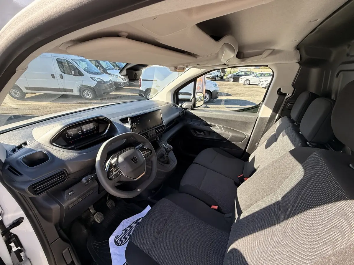 Vue intérieure côté conducteur du Peugeot Partner blanc 2026, montrant le tableau de bord et les sièges avant en tissu noir.