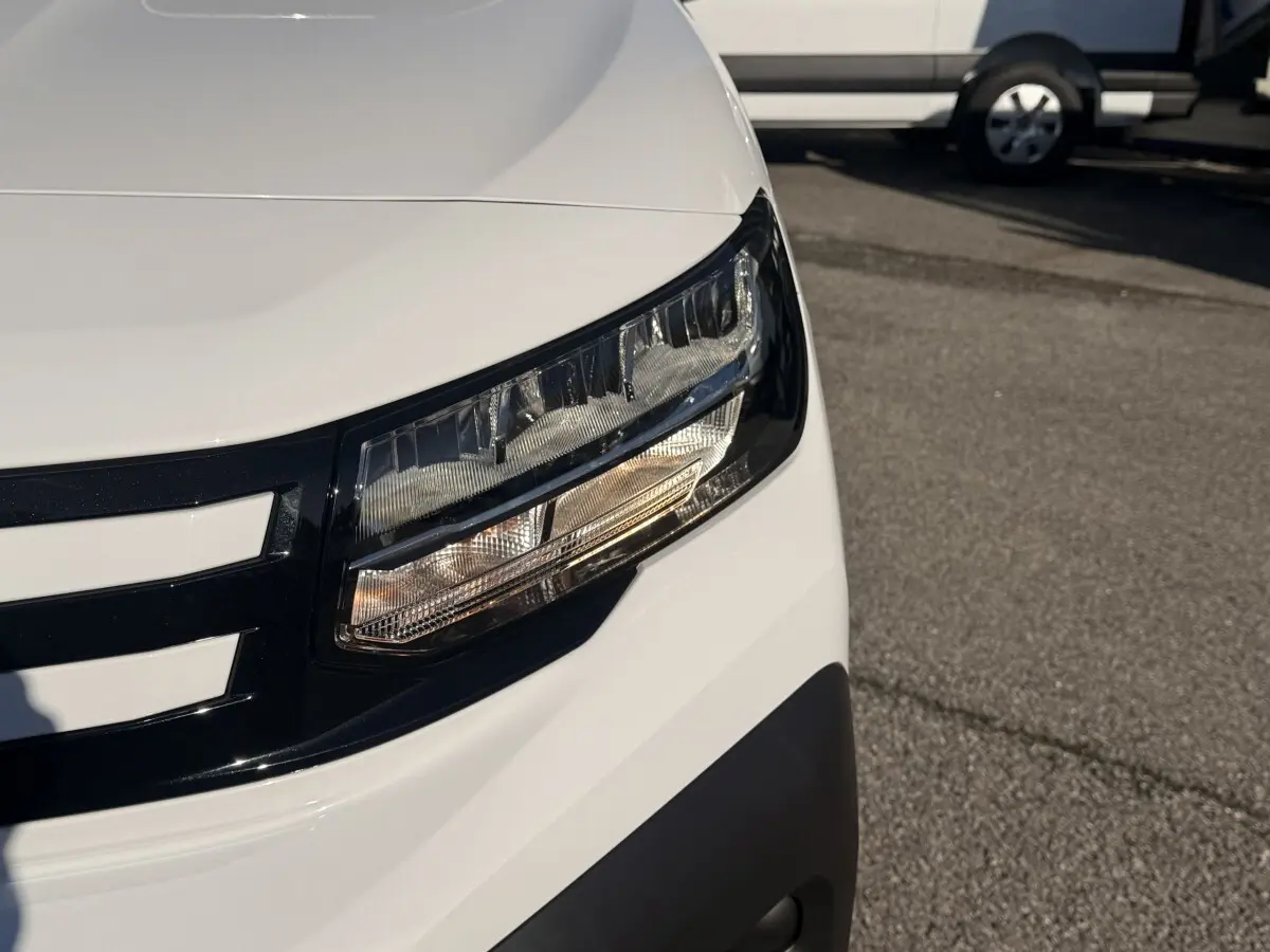 Gros plan sur le phare avant droit du Peugeot Partner blanc, soulignant la calandre noire et le pare-chocs gris.