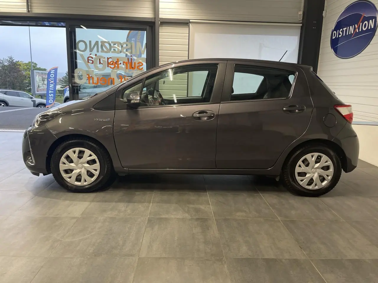 Profil côté gauche d'une Toyota Yaris hybride 2020 gris foncé métallisé avec enjoliveurs blancs en intérieur showroom.