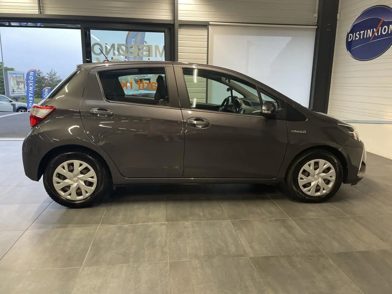 Profil côté gauche d'une Toyota Yaris hybride gris foncé métallisé, avec enjoliveurs 15 pouces et calandre noir laqué visible en showroom.