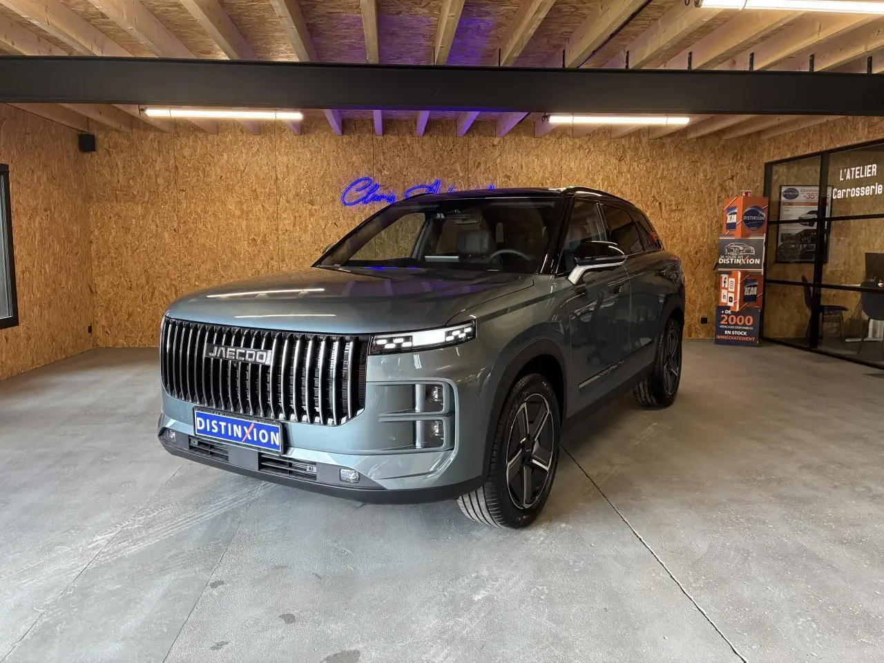 SUV JAECOO J7 gris olive en 3/4 avant droit, avec toit noir et calandre verticale imposante, en intérieur showroom.