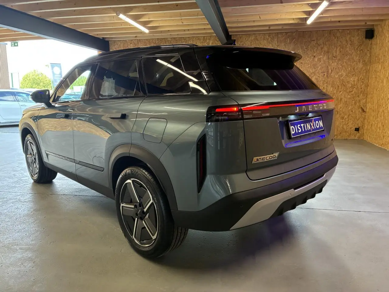 SUV JAECOO J7 gris olive vu en 3/4 arrière droit, toit noir et feux arrière LED allumés dans un garage.