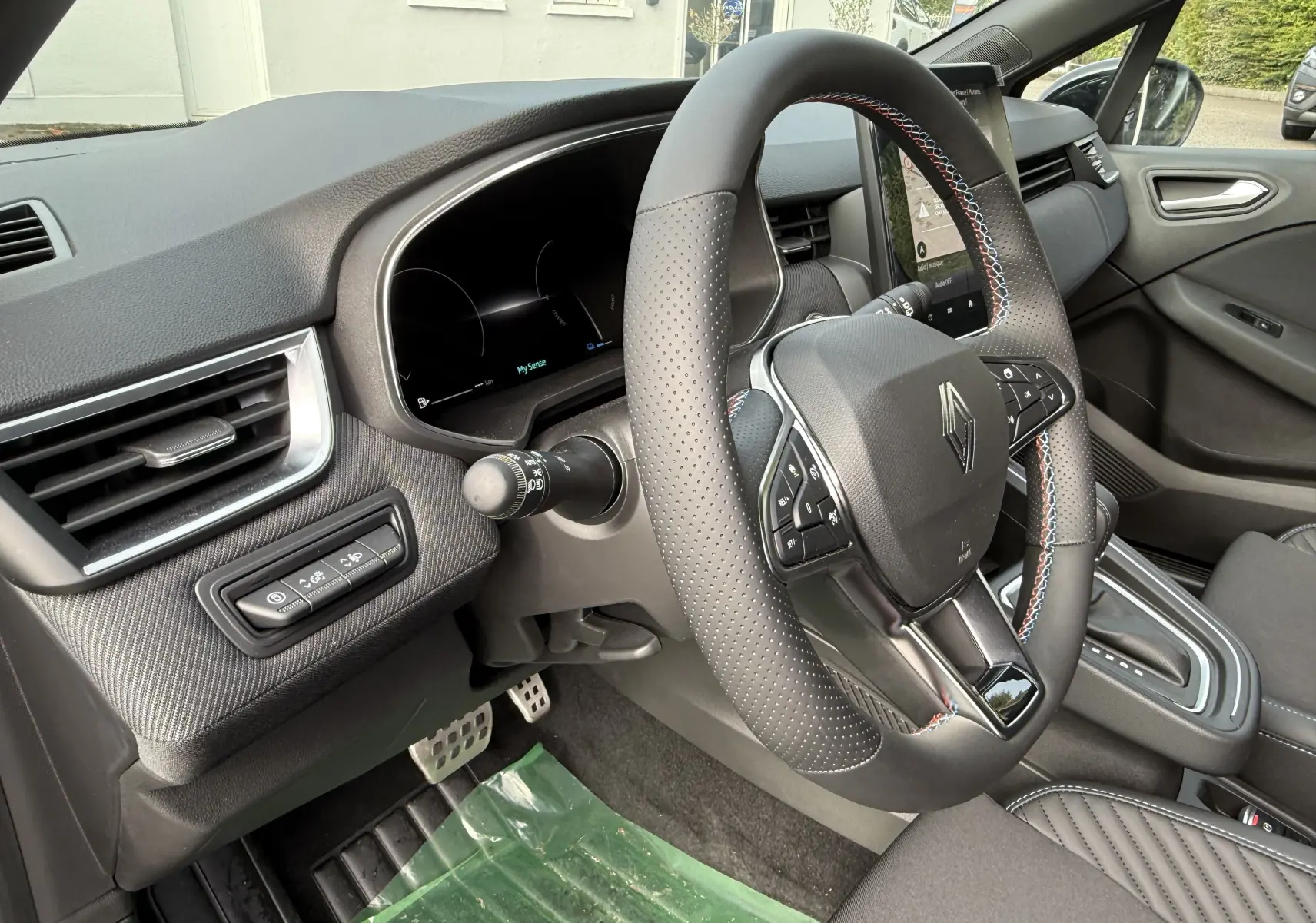 Vue intérieure côté conducteur de la Renault Clio V hybride 2025, volant cuir perforé avec surpiqûres tricolores et tableau de bord numérique.