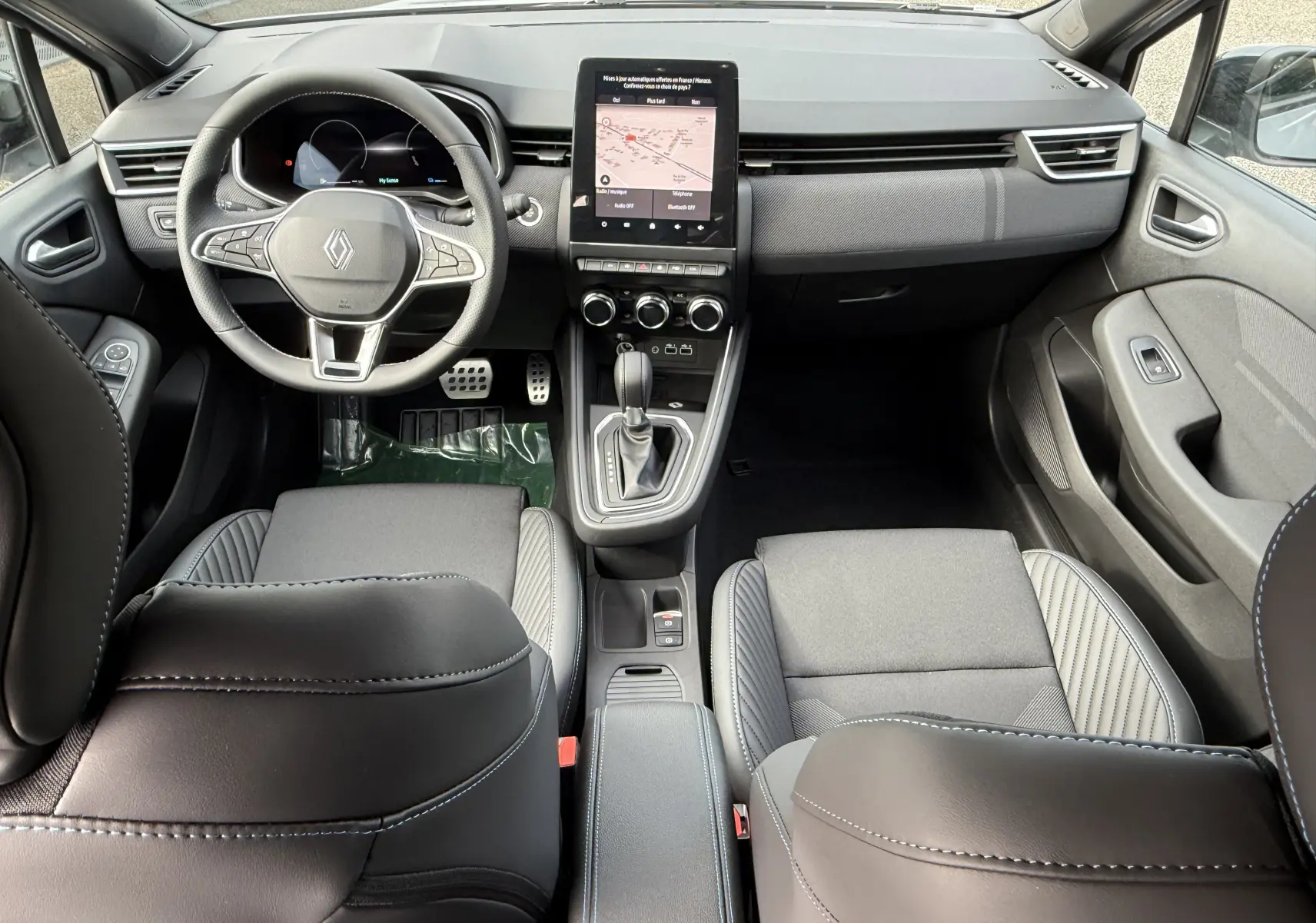 Vue intérieure avant de la Renault Clio V 1.6 E-TECH avec tableau de bord moderne, écran tactile vertical et sièges noirs.