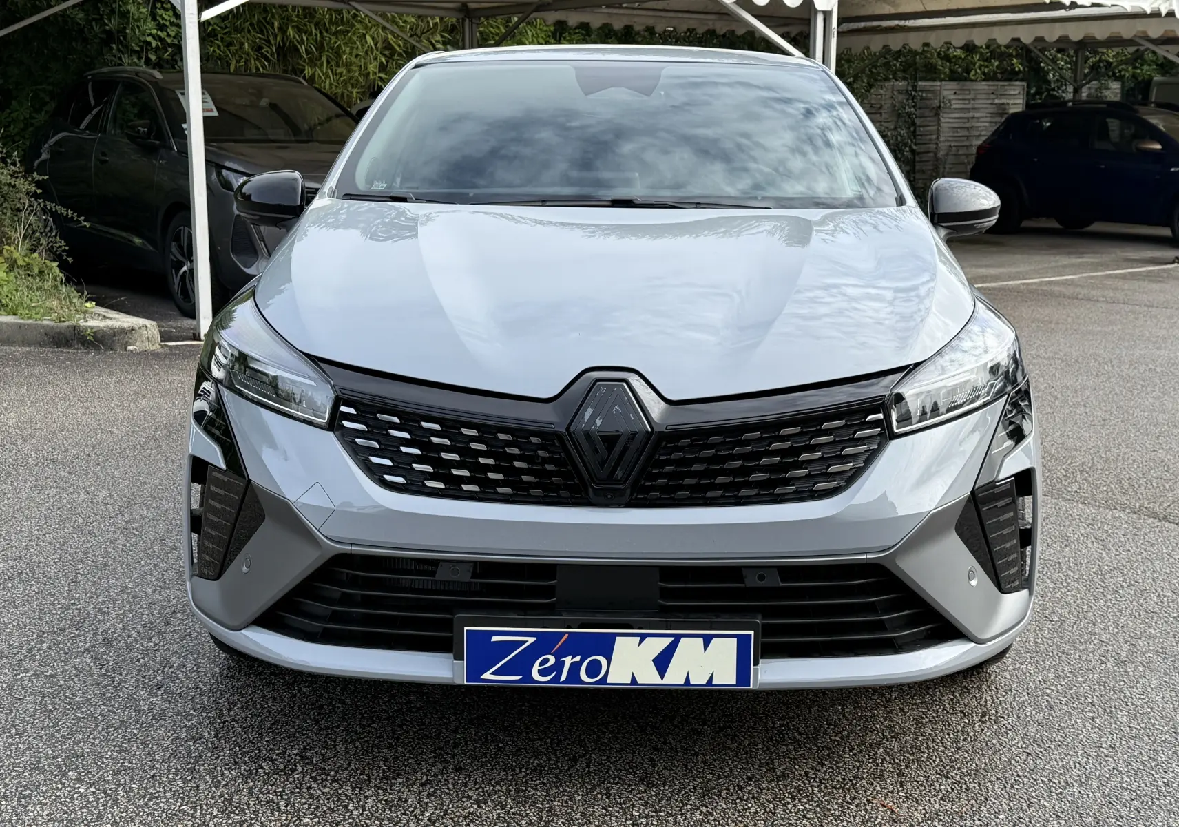 Vue frontale d'une Renault Clio V gris rafale avec calandre noire et plaque d'immatriculation "Zéro KM" visible.
