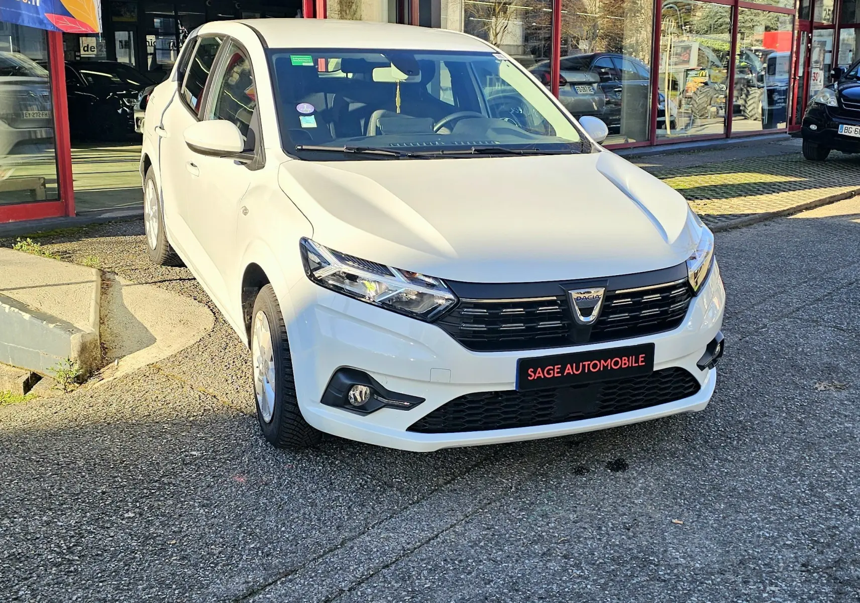 Vue 3/4 avant d'une Dacia Sandero III blanche glacier, avec calandre noire et phares LED, stationnée devant un showroom.