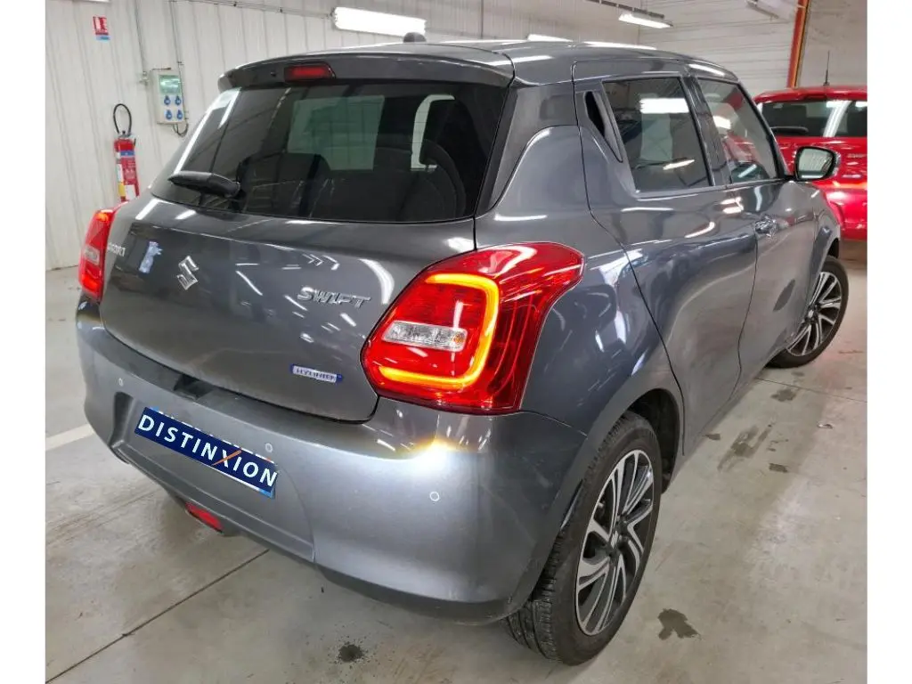 Vue 3/4 arrière droite d'une Suzuki Swift 1.2 Dualjet Hybrid grise de 2023 avec feux arrière allumés en intérieur.
