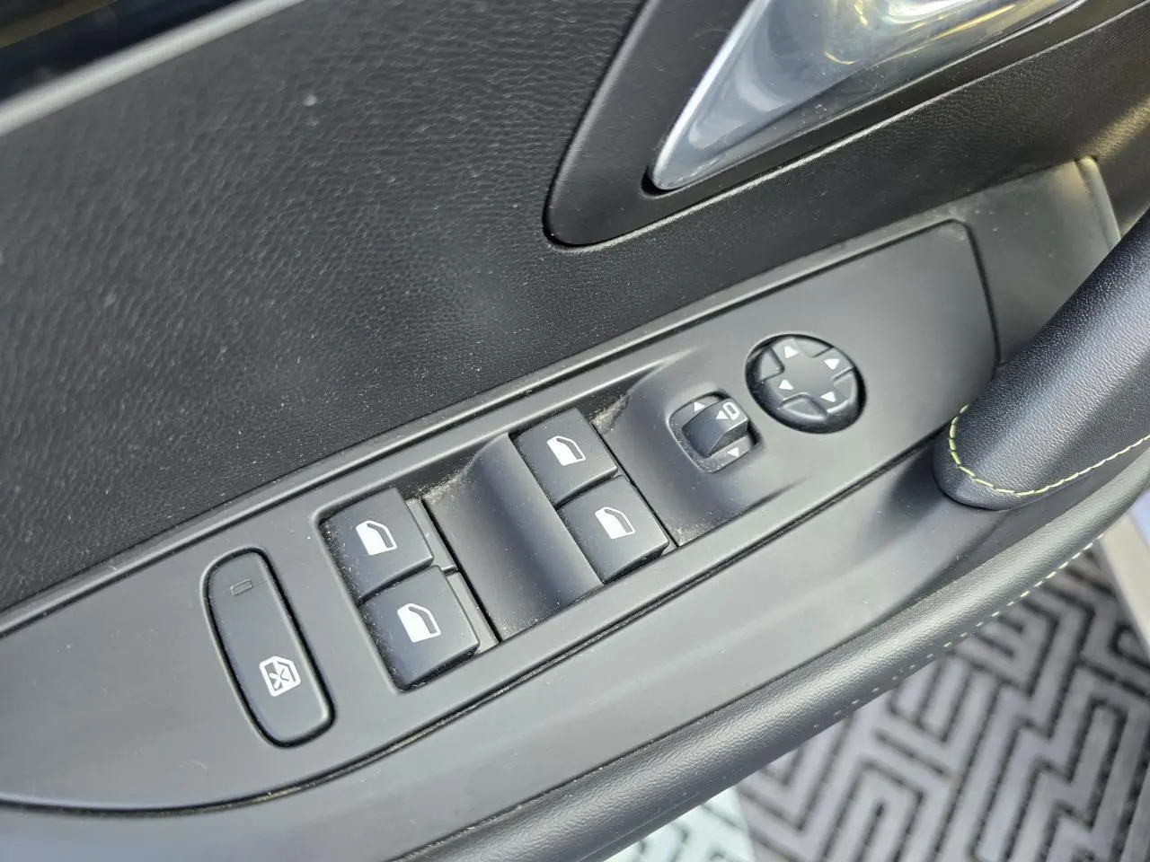 Gros plan sur les commandes de lève-vitres et rétroviseurs côté conducteur de la Peugeot 208 grise, avec surpiqûres vertes visibles.