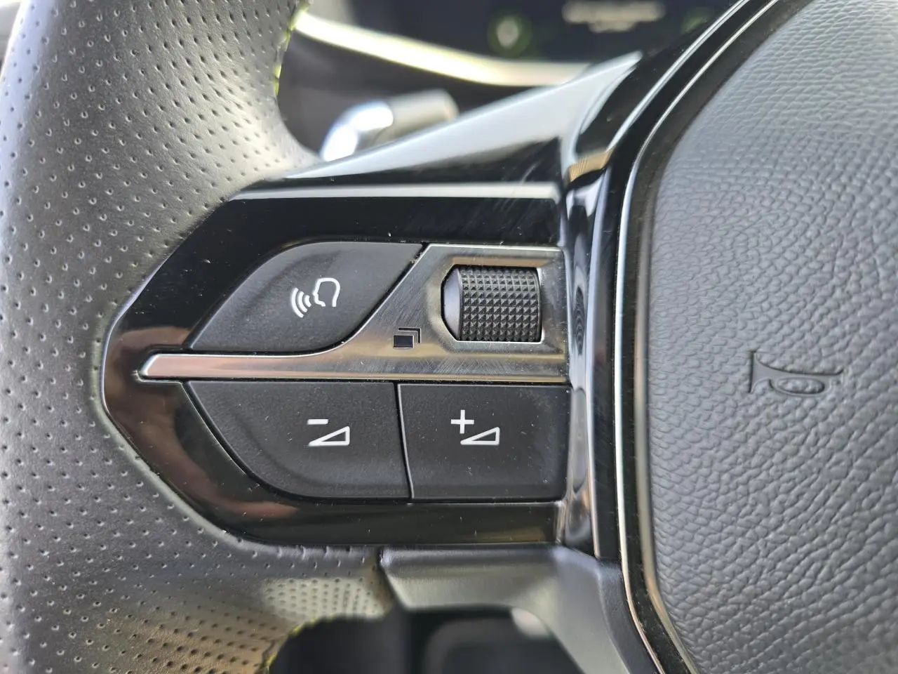 Gros plan sur les commandes de volume et reconnaissance vocale sur le volant noir perforé d'une Peugeot 208 grise 2023.