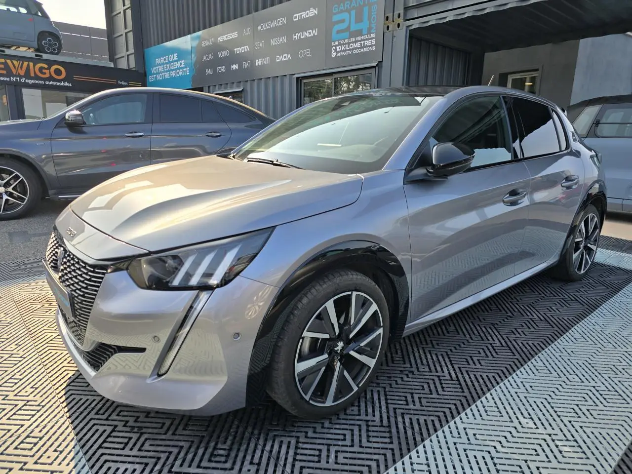 Peugeot 208 grise vue 3/4 avant droit avec jantes alliage et toit noir brillant dans un showroom extérieur.