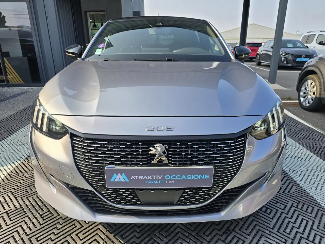 Vue frontale d'une Peugeot 208 grise 2023 avec calandre noire et feux LED à signature 3 griffes.