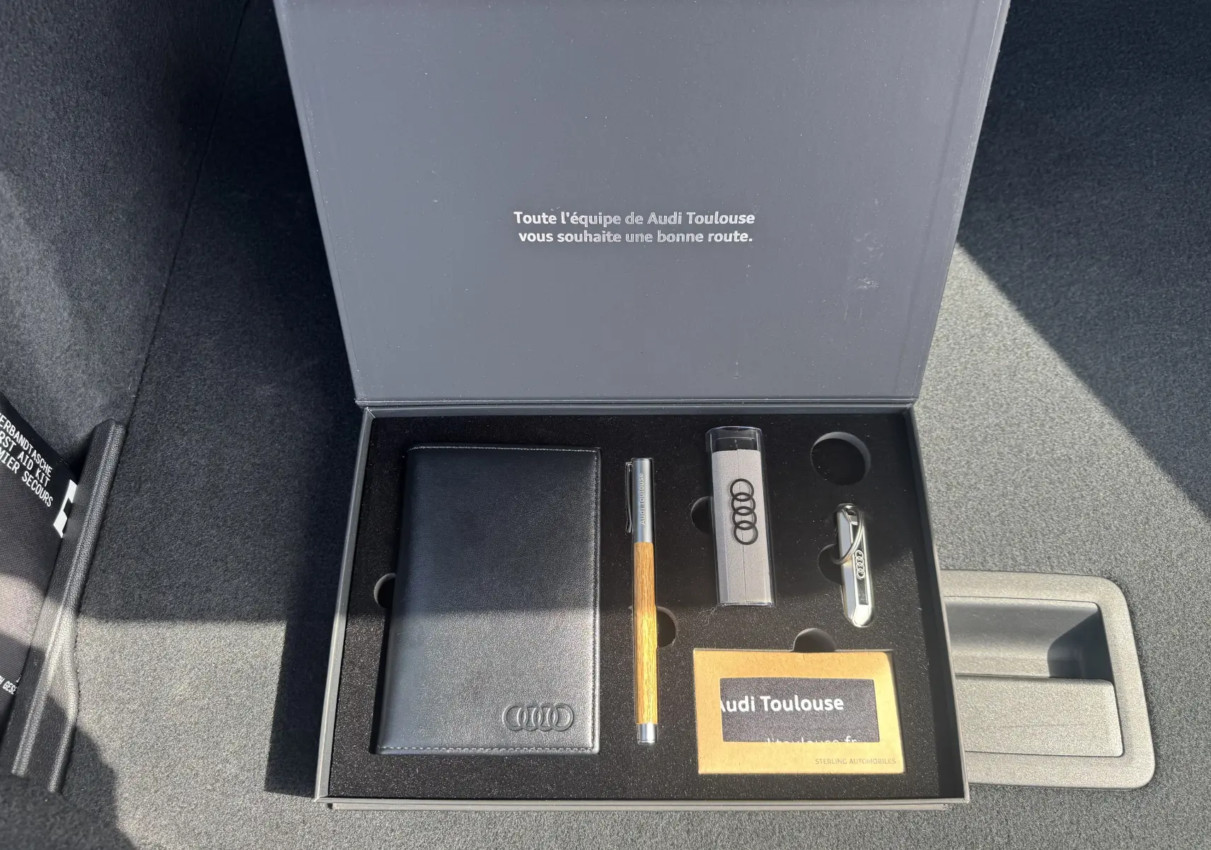 Boîte ouverte dans le coffre d’une Audi A3 Sportback gris, contenant porte-documents, stylo et clés Audi.