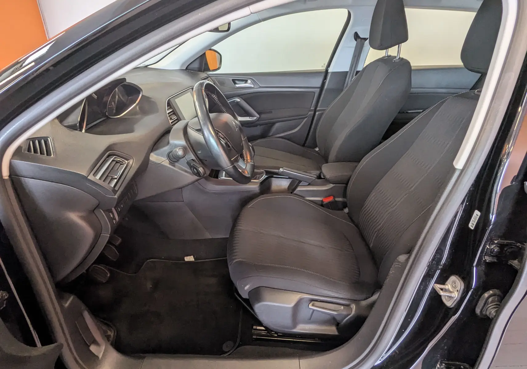 Vue intérieure côté conducteur de la Peugeot 308 noire de 2017, montrant les sièges en tissu gris et le tableau de bord.
